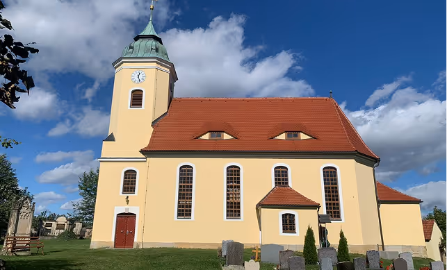 Gelbe Kirche mit rotem Ziegeldach und markanten Dachfenstern, umgeben von einem Friedhof mit Grabsteinen unter blauem Himmel.