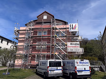 Renoviertes Gebäude in Rosa mit Gerüst und zwei weißen Wohütt-Maler- und Bodenlegerfahrzeugen davor.