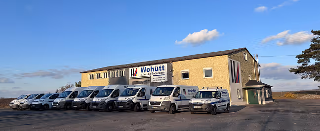 Neun weiße Fahrzeuge der Firma Wohütt vor einem großen, hellen Firmengebäude unter blauem Himmel.