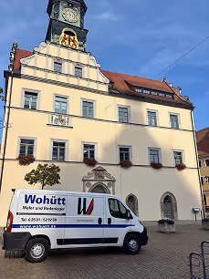 Weißer Transporter mit Werbung 'Wohütt Maler und Bodenleger' steht vor einem historischen Gebäude mit Uhrenturm.