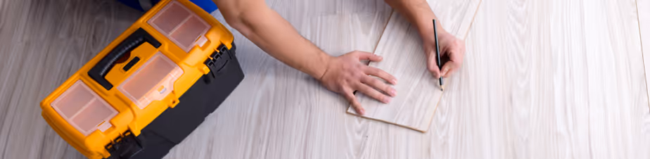 Person hält eine kleine Holzplatte auf einem hellen Holzfußboden und markiert eine Stelle mit einem Bleistift neben einem orange-schwarzen Werkzeugkasten.