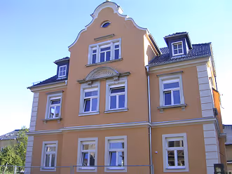 Hellgelbes historisches Wohngebäude mit vielen Fenstern und dekorativem Giebel vor blauem Himmel.