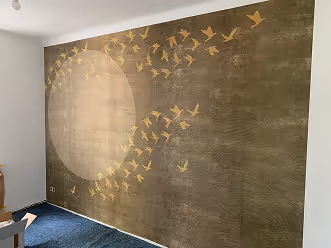 Raum mit einer hellen Wand, verziert mit einem großen, hellen Kreis und goldfarbenen Vogelsilhouetten, die nach rechts fliegen.
