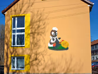 Hauswand mit zwei Fenstern, die von gelben Rahmen umgeben sind, und einer Wandmalerei eines niedlichen Bären mit Kochmütze, der an einem grünen Untergrund sitzt.